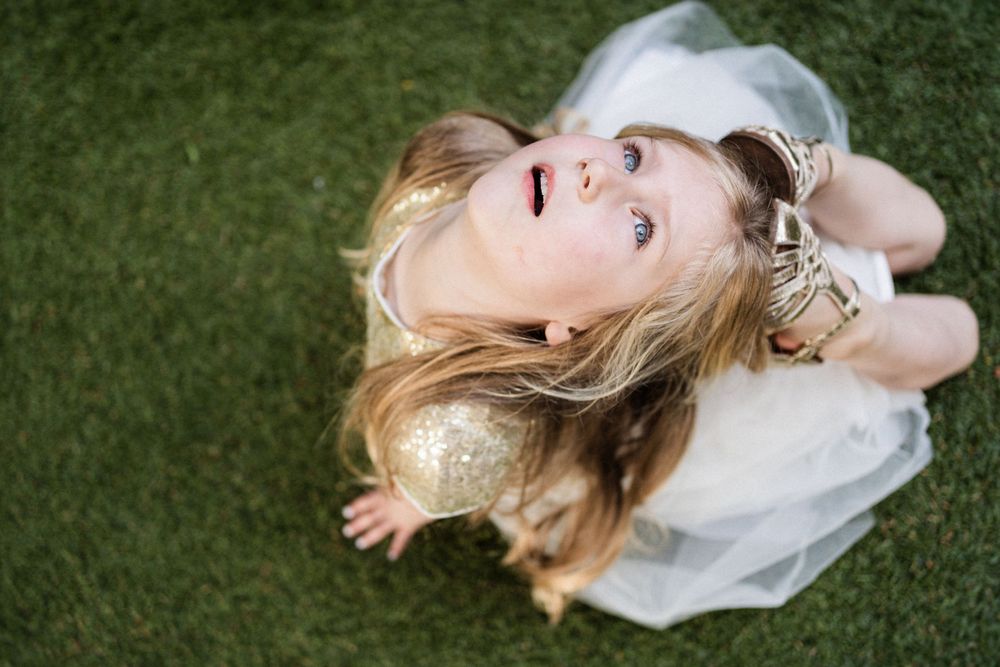 A young person with blonde hair lies on green grass wearing a white dress looking up at the camera.