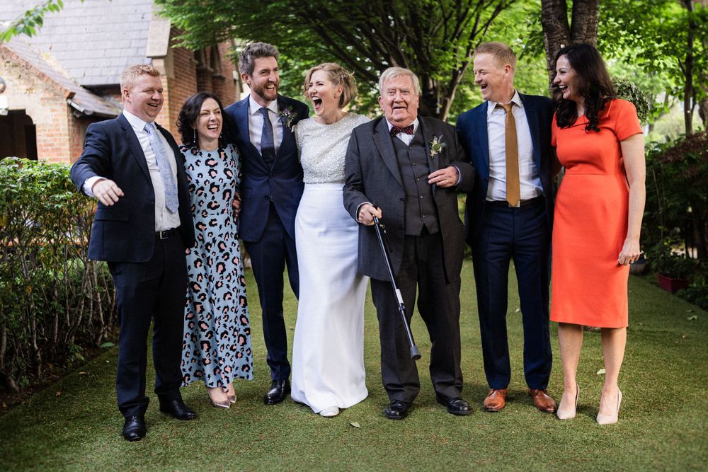 A group of wedding guests pose together outdoors in formal attire on a sunny day.