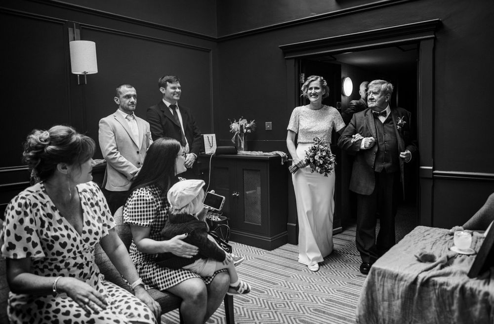A black and white candid photo of wedding guests gathered in a dimly lit room during a ceremony.