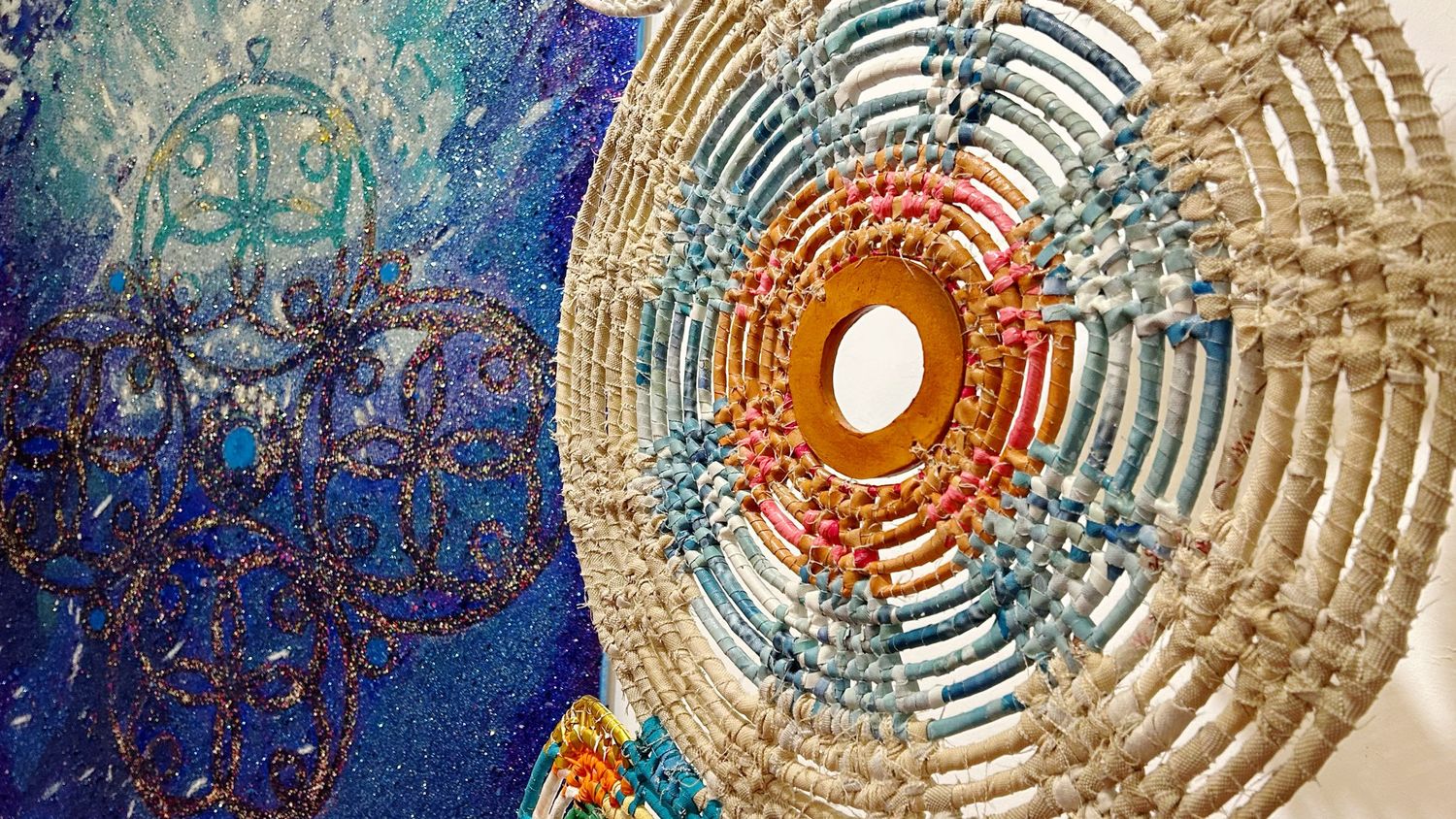 Close-up of circular woven artwork next to blue abstract painting in gallery setting.