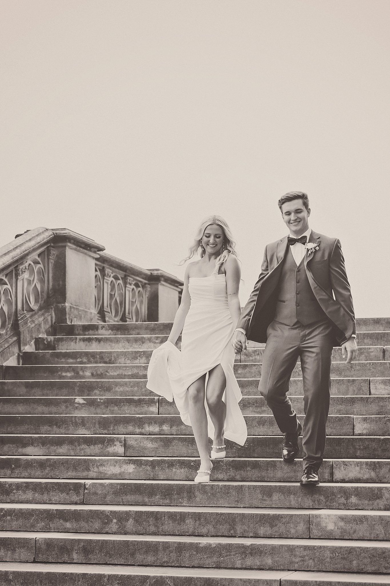 Black and white photo of couple descending ornate stone staircase in wedding attire.