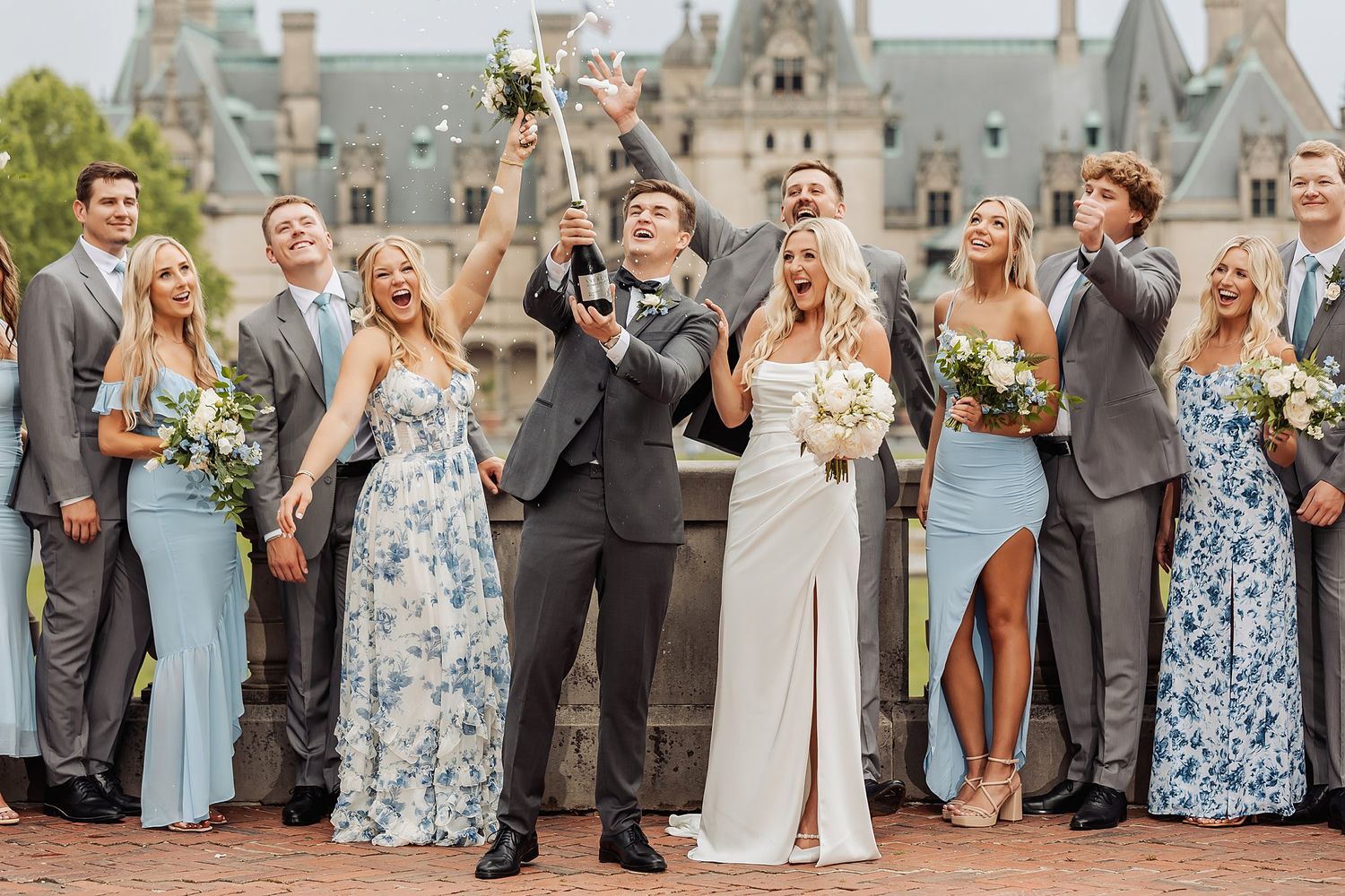 Wedding party celebrates with champagne in front of elegant castle backdrop.
