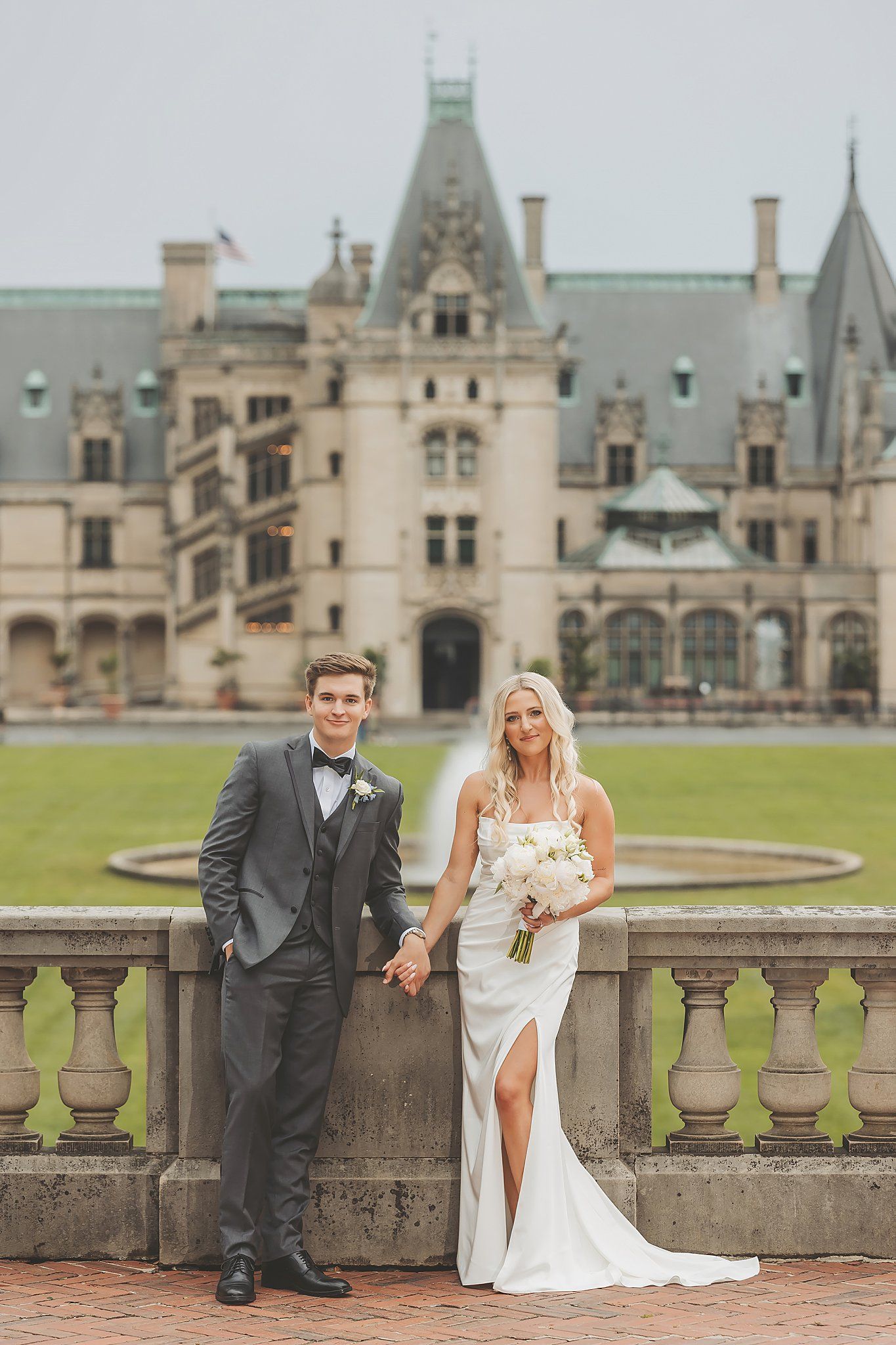 Elegant wedding couple poses on stone balcony with majestic Biltmore Estate in background.