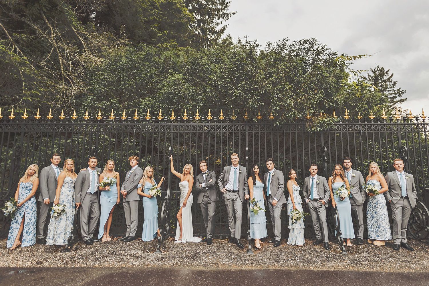 Wedding party in light blue and gray attire poses against an ornate iron fence outdoors.