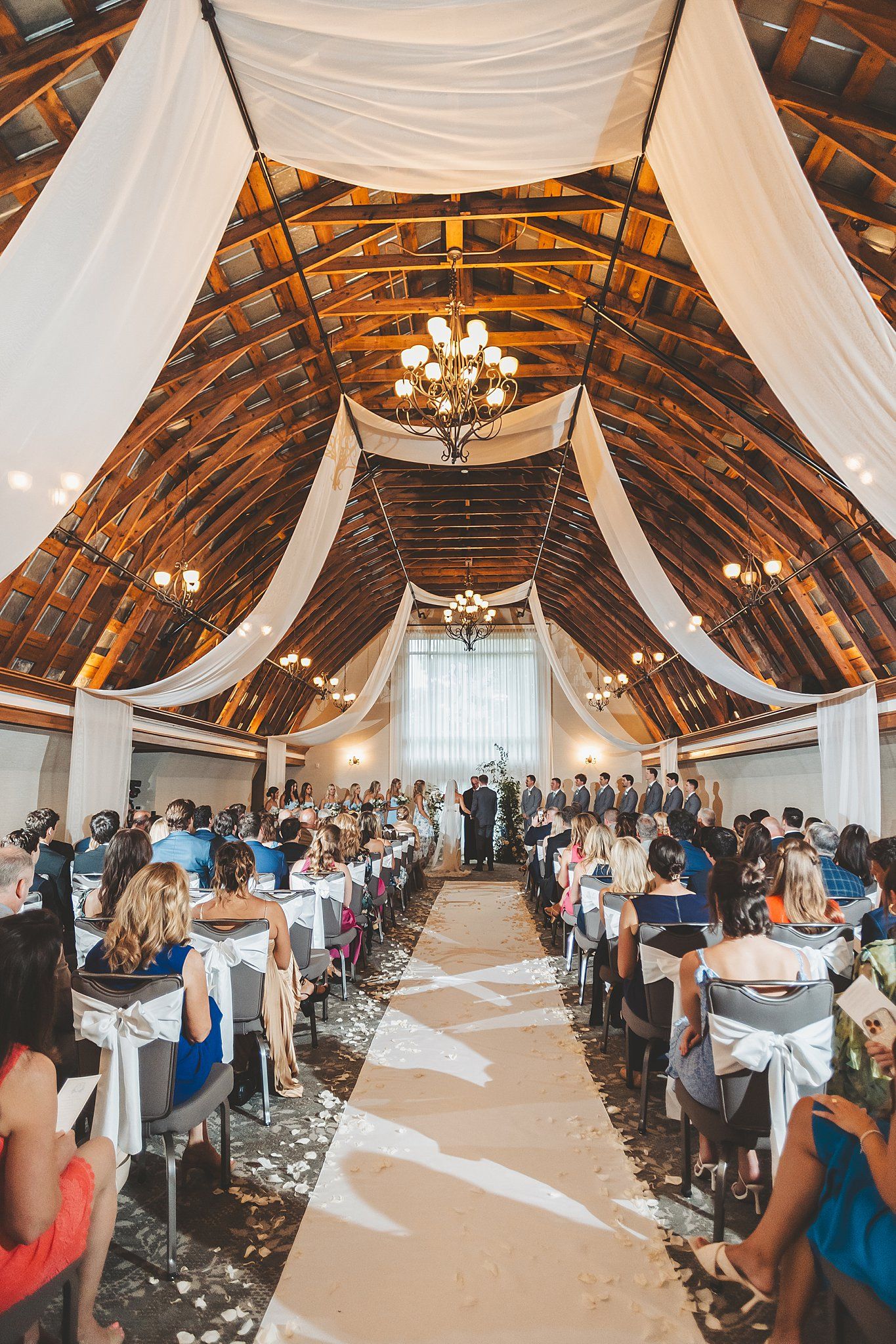 Elegant barn wedding ceremony with white draping and chandeliers creating a dramatic ceiling.