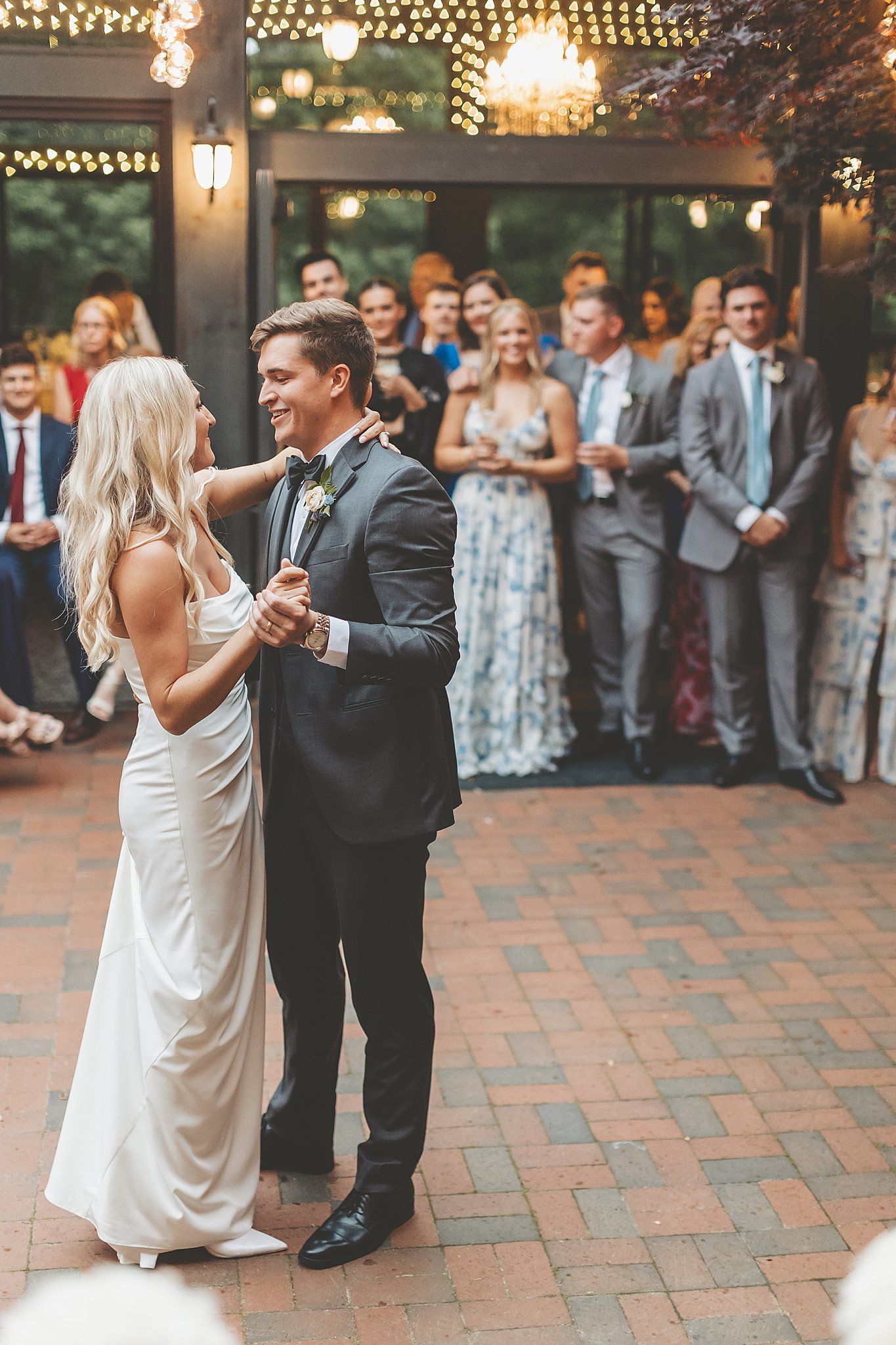 First dance on brick patio with wedding guests watching in background.