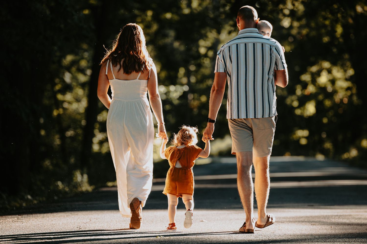 Family walking together on a sunlit path with a small child in orange clothing between them.