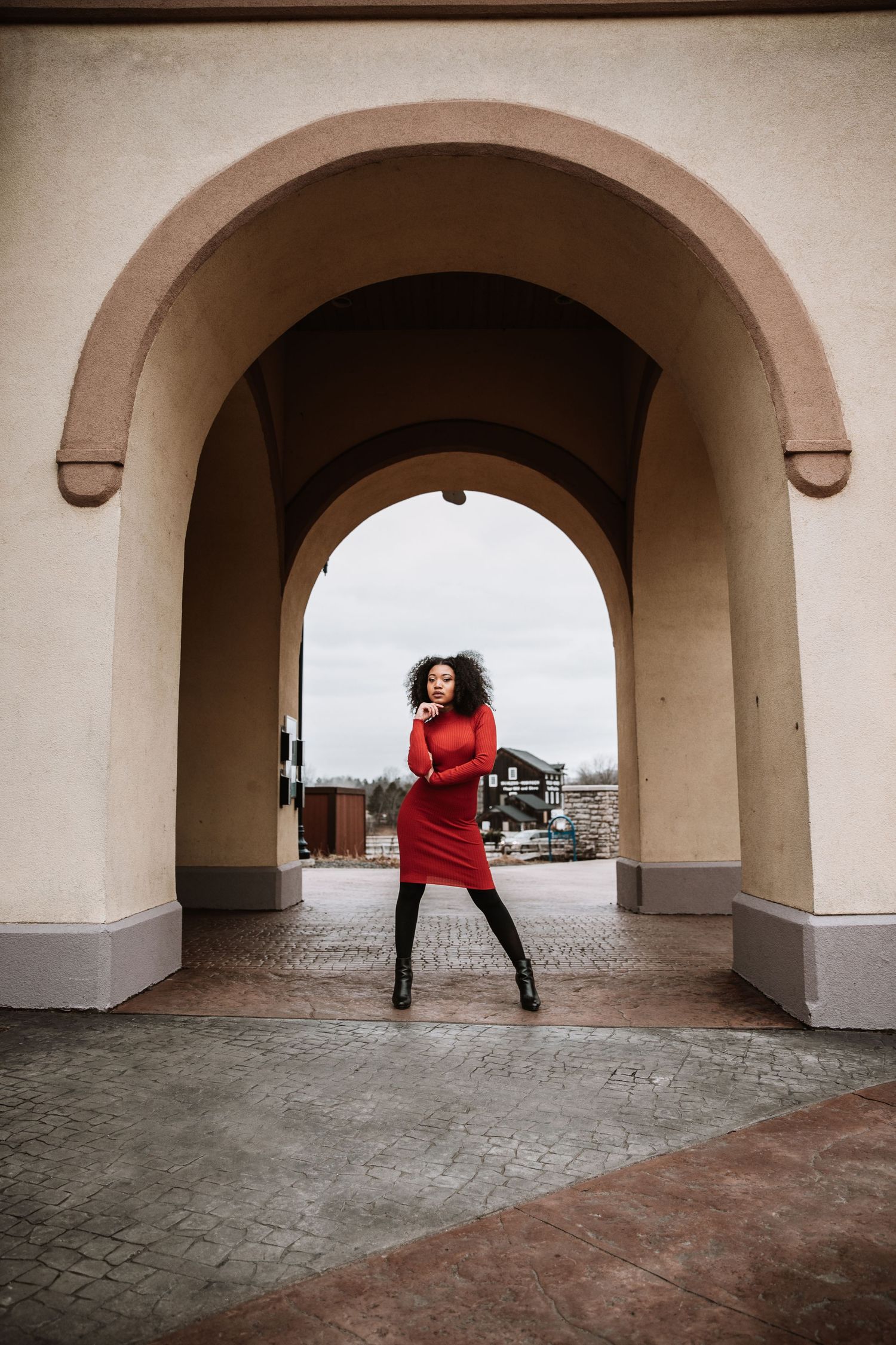 Model in red dress poses dramatically under stone archway in urban setting.