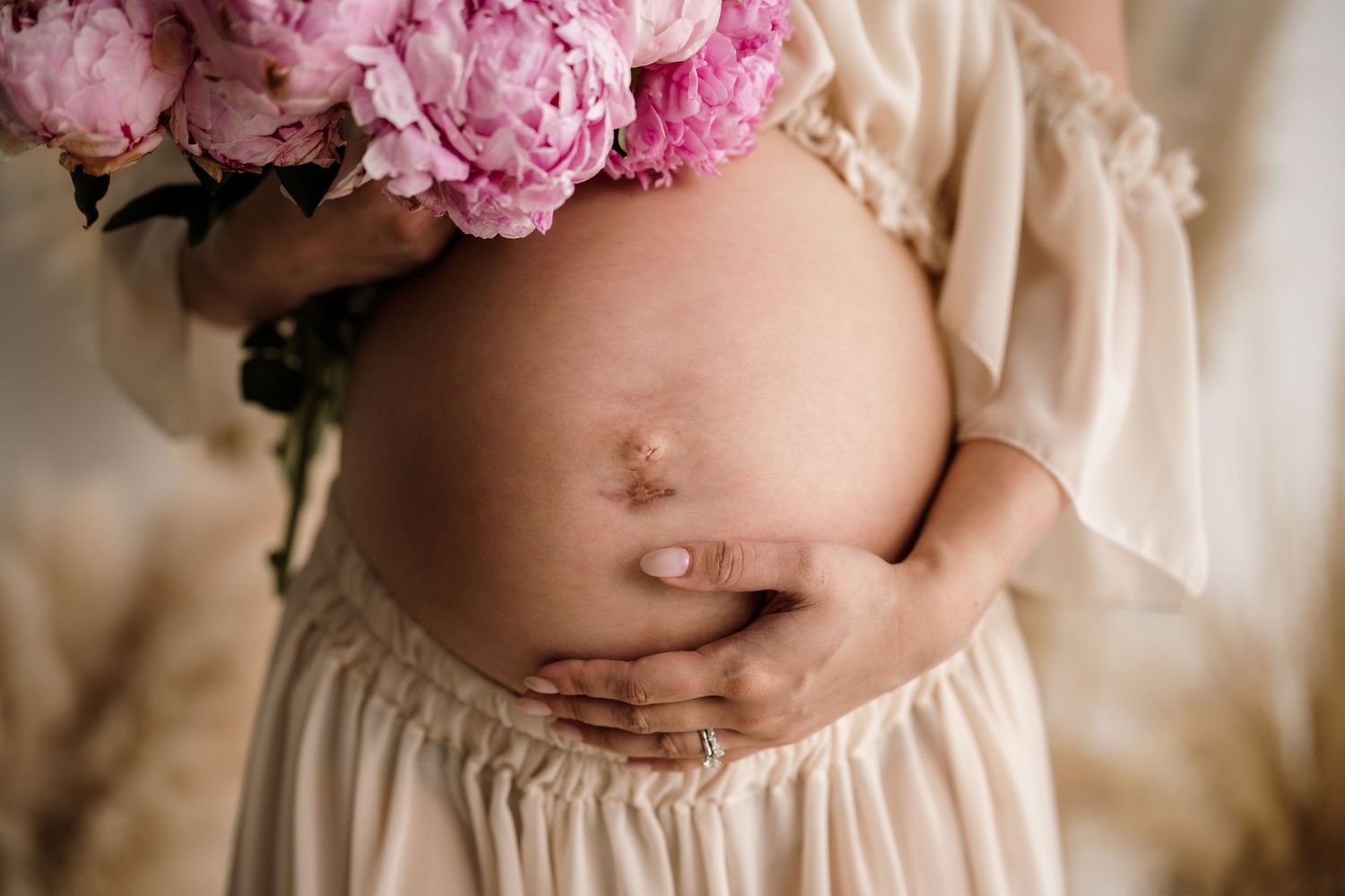 Close up of hands making heart shape around pregnant belly in cream colored dress.