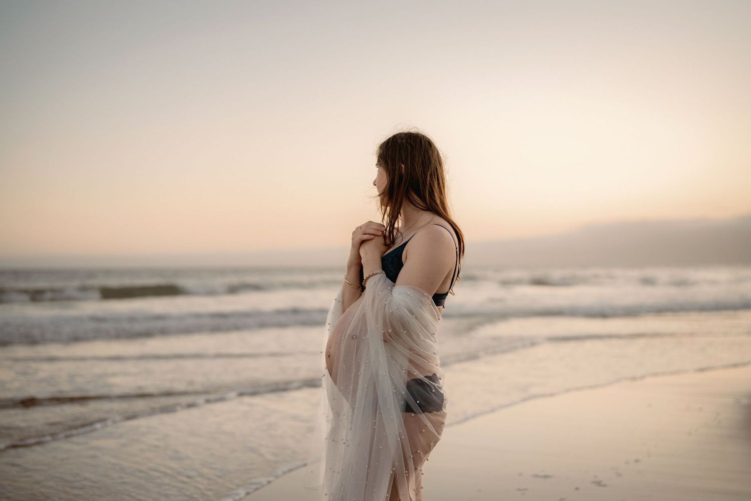 Close up of hands making heart shape around pregnant belly in cream colored dress.