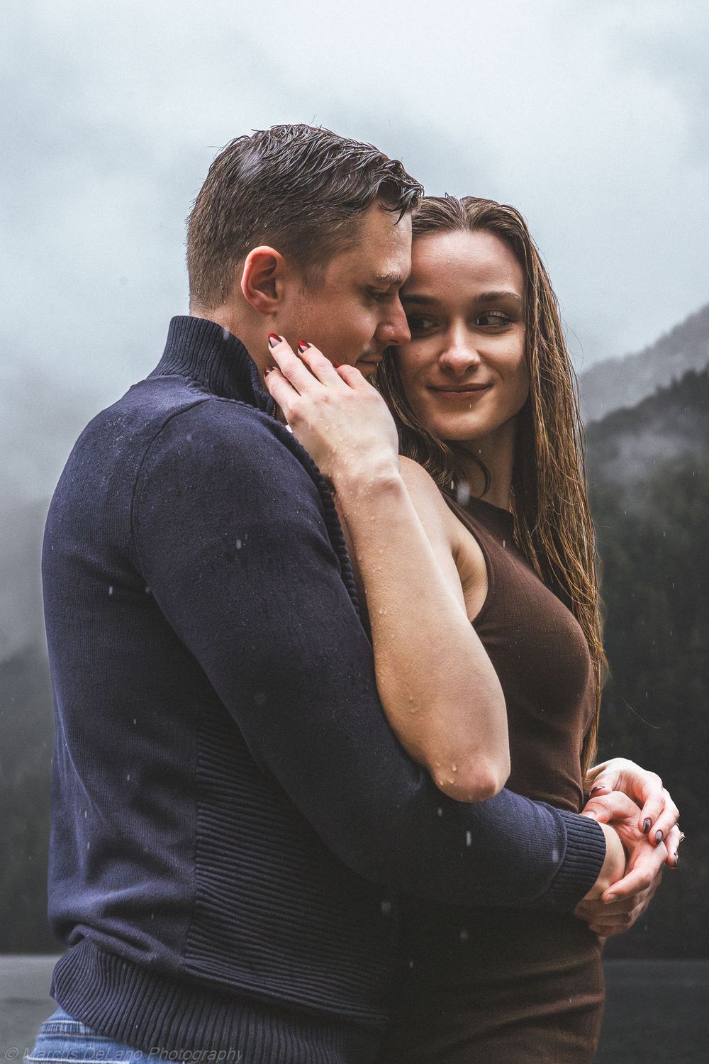 Romantic couple embraces outdoors on cloudy day with mountains in background.