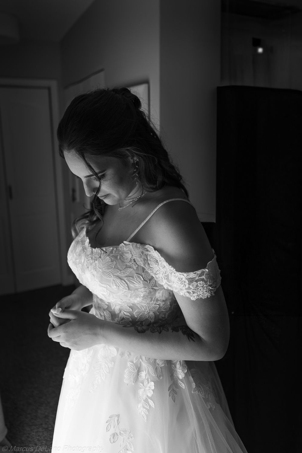 A black and white portrait of someone in an off-shoulder lace wedding dress standing in soft lighting.