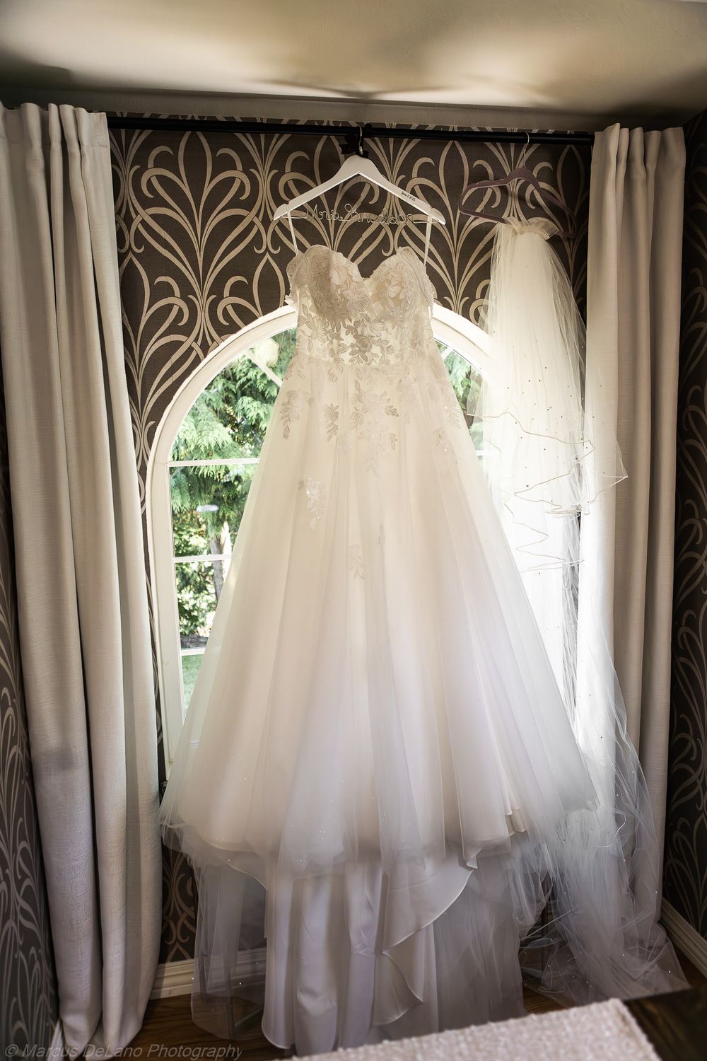 Four identical white wedding dresses with tulle skirts hang from windows against a dark patterned wallpaper background.