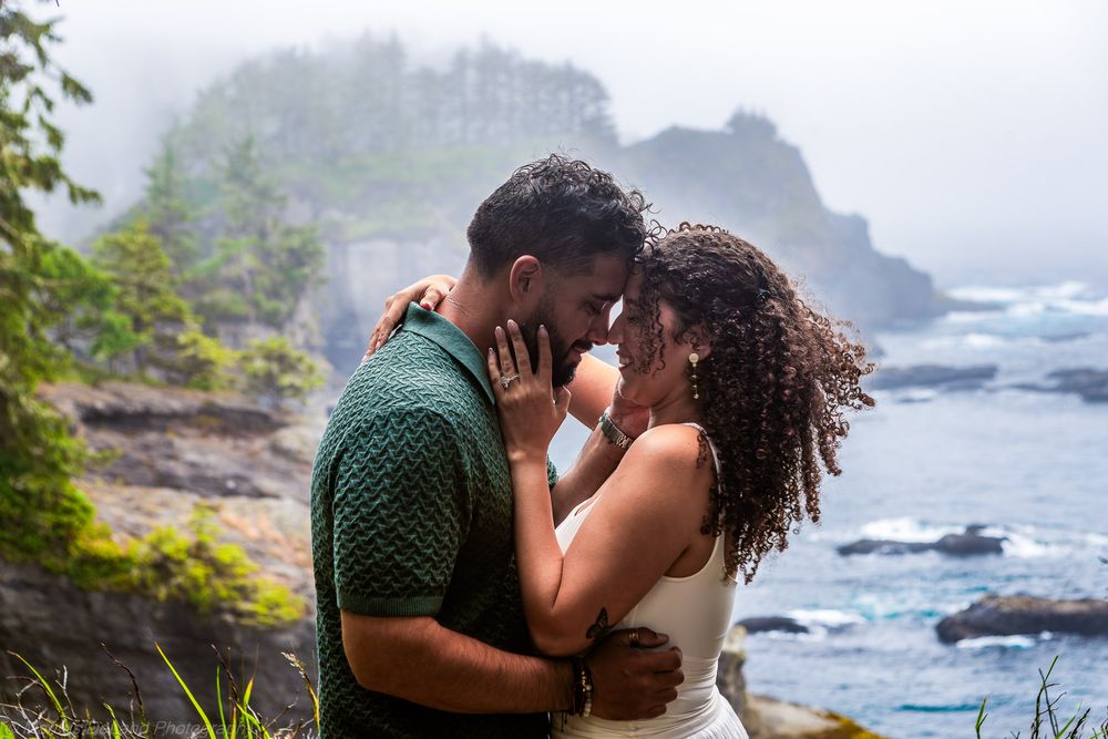 Romantic couple embraces on foggy Pacific Northwest coastline with rocky cliffs and ocean waves in background.