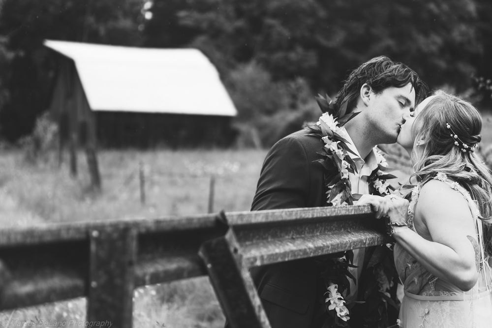 Black and white photo of a couple sharing a kiss at their outdoor wedding ceremony near a rustic fence and barn.