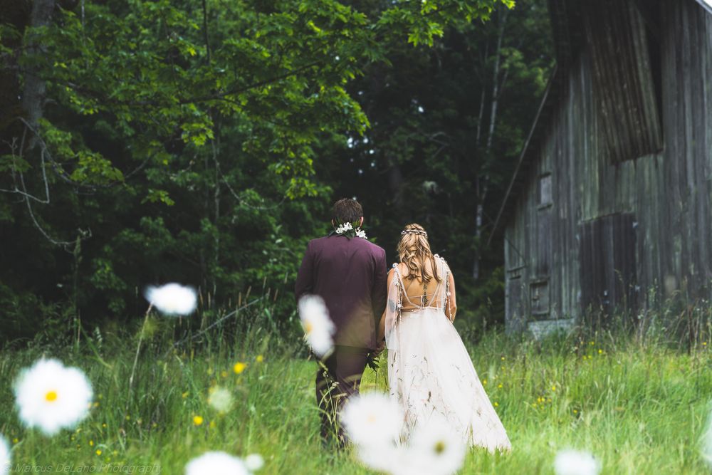 Bride and groom walking through a wildflower meadow toward a rustic barn on their wedding day.