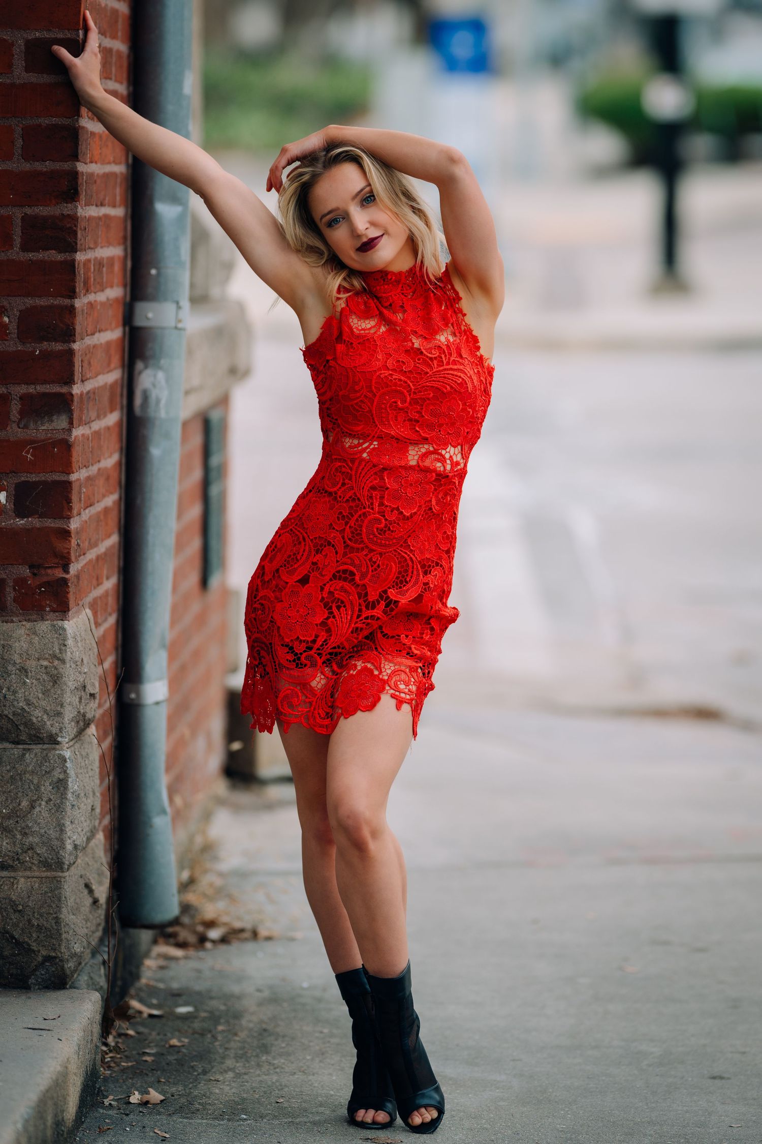 A person in a vibrant red lace dress poses playfully against a brick wall in an urban setting.