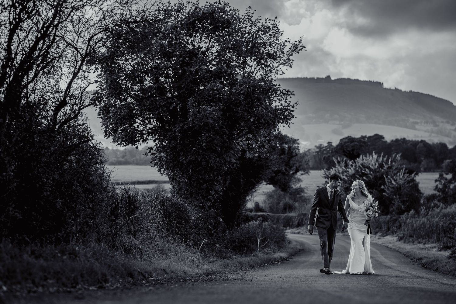 A couple walks down a scenic country road with mountains and trees silhouetted against a dramatic cloudy sky.