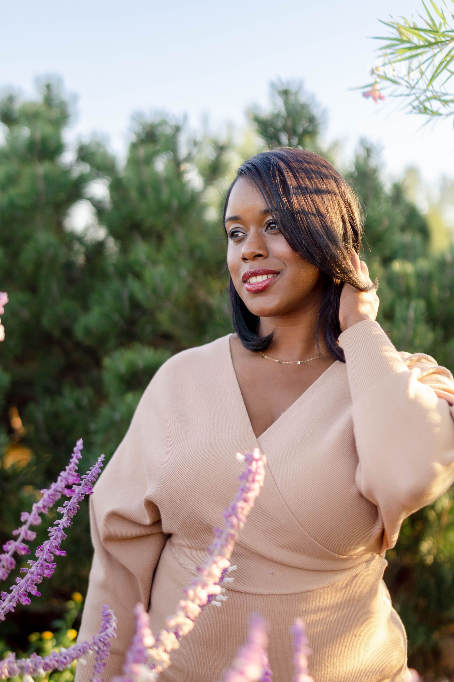 Elegant Black woman in beige wrap dress posing among purple lavender flowers and pine trees.