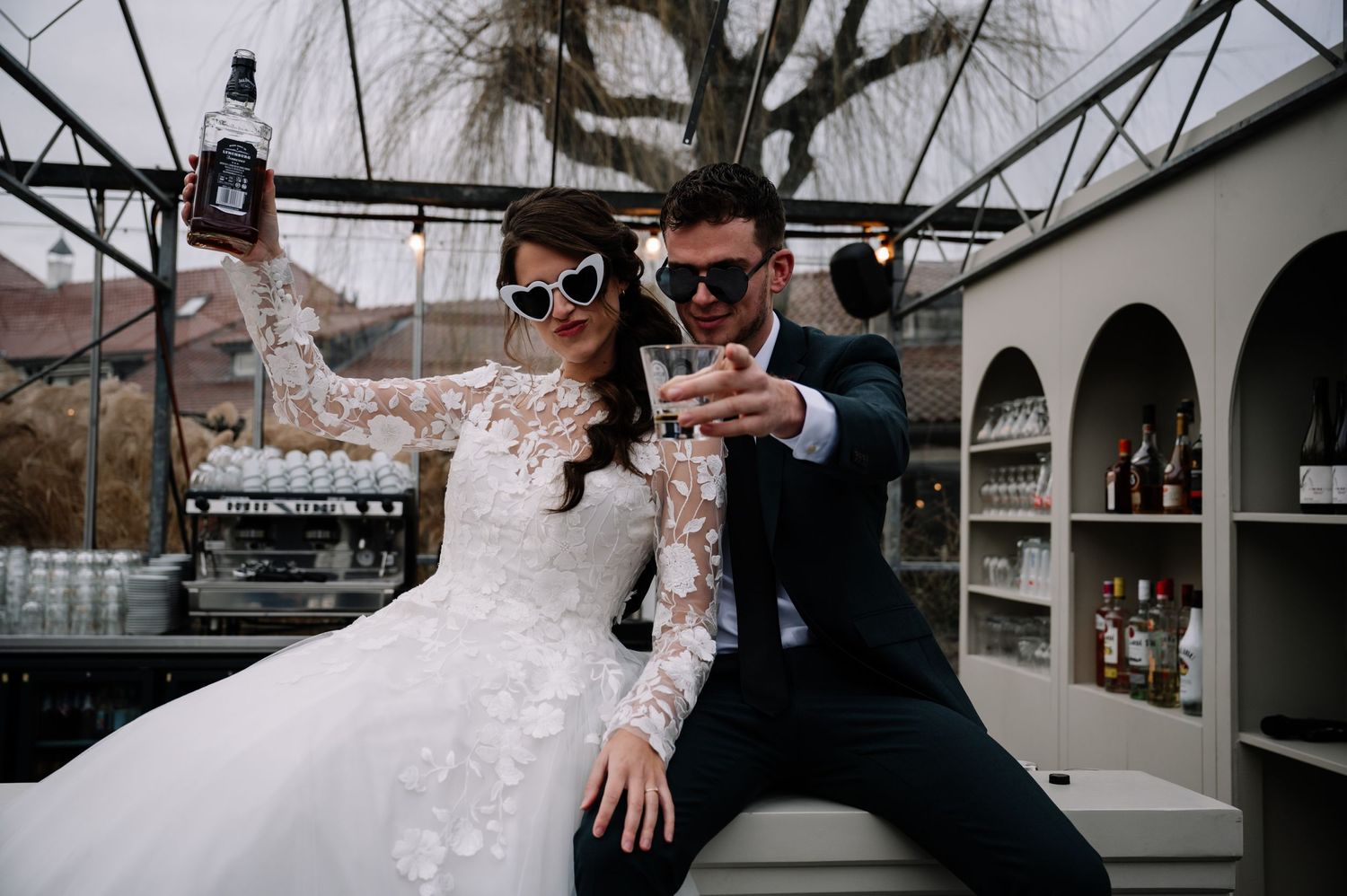 A couple wearing sunglasses poses with drinks at an outdoor bar during their wedding celebration.