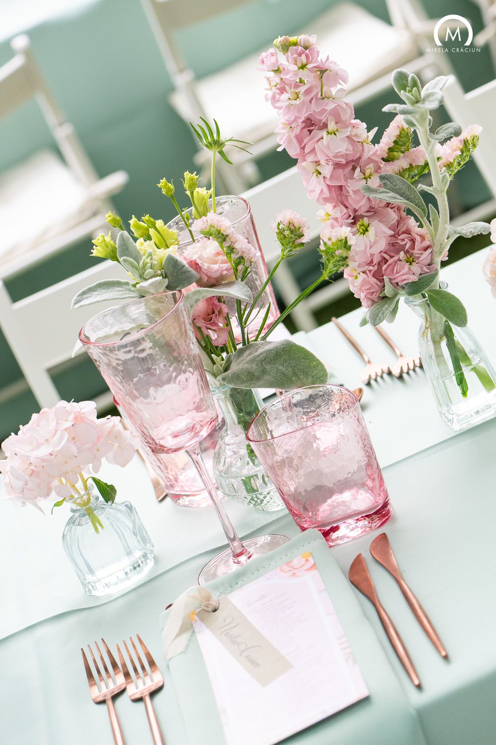 Elegant table setting with pink flowers in glass vases, rose-tinted glassware, and copper utensils on mint green tablecloth.
