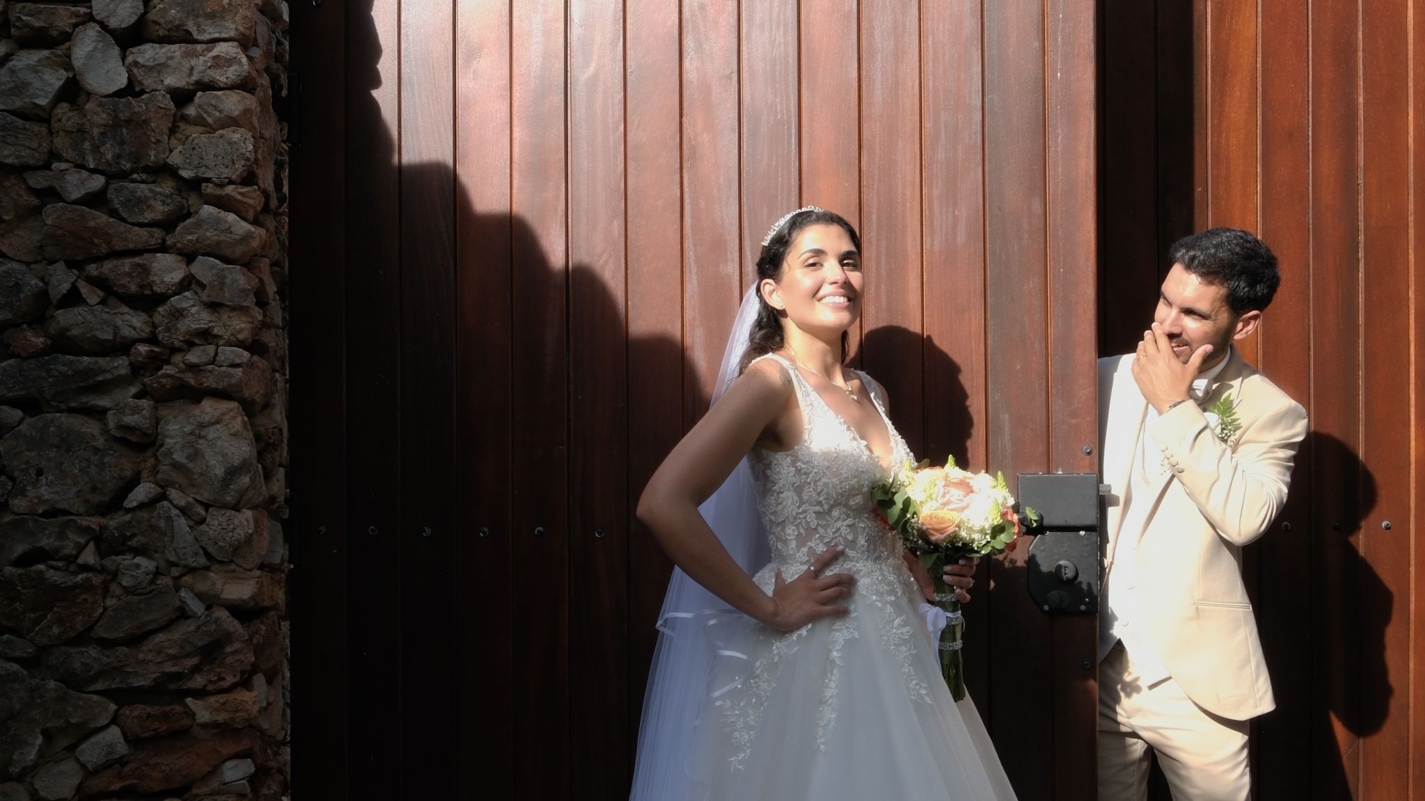 Smiling bride and groom standing beside a large wooden gate on their wedding day, at "As Fontes Events Venue" in Alte, Algarve.
