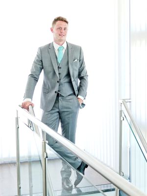 Professional in a light gray three-piece suit with turquoise tie stands confidently on modern staircase with glass railing.
