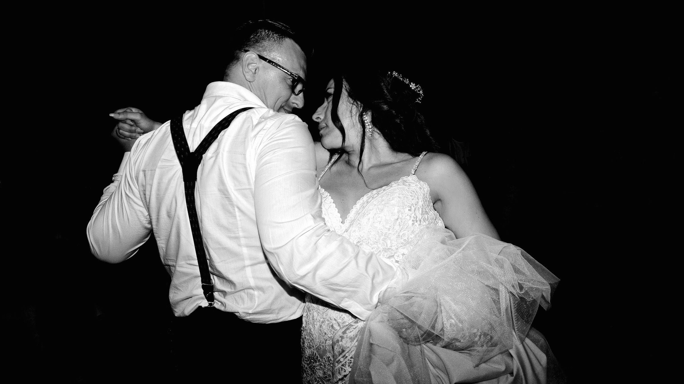 Black and white photo of a couple dancing together at a wedding reception with the groom wearing suspenders.