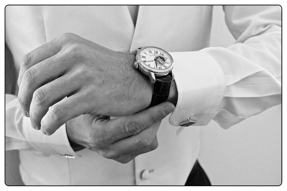 Black and white close-up photograph of hands adjusting a luxury watch on a suit sleeve.