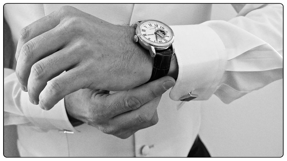 Black and white close-up photograph of hands adjusting a luxury watch on a suit sleeve.