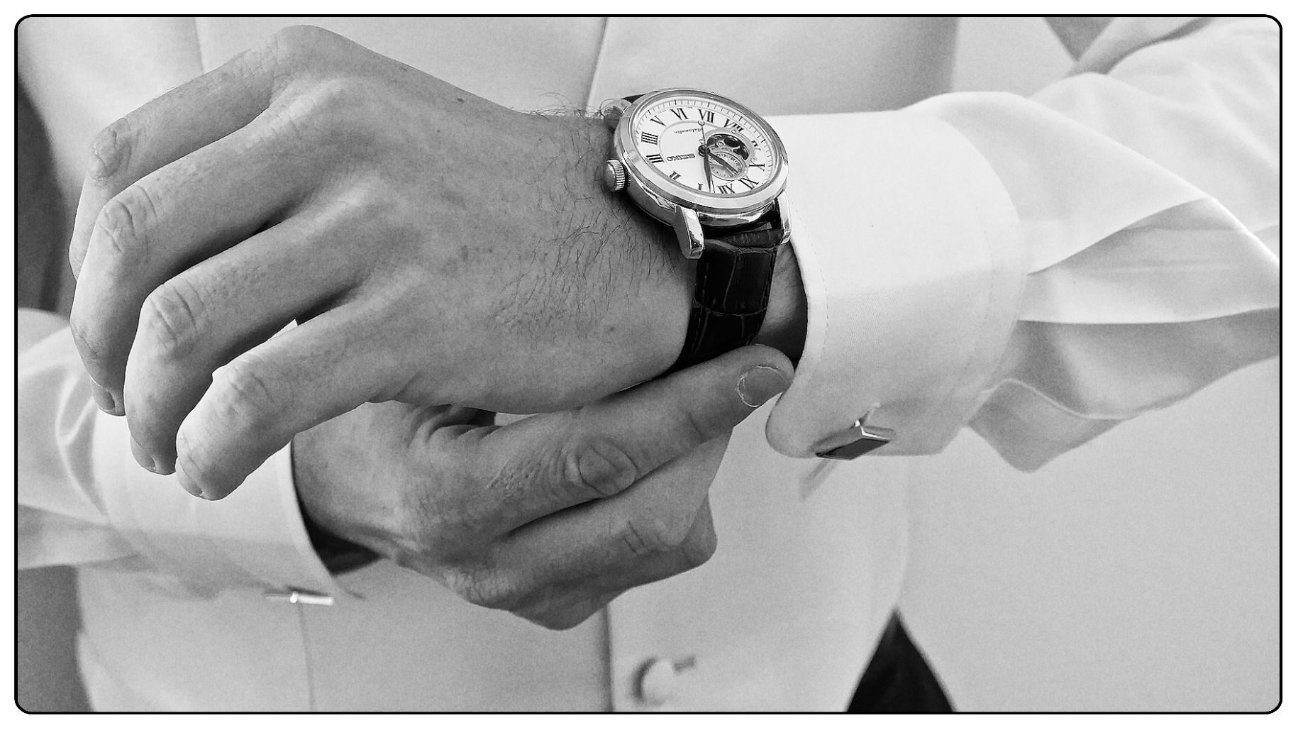 Black and white close-up photograph of hands adjusting a luxury watch on a suit sleeve.