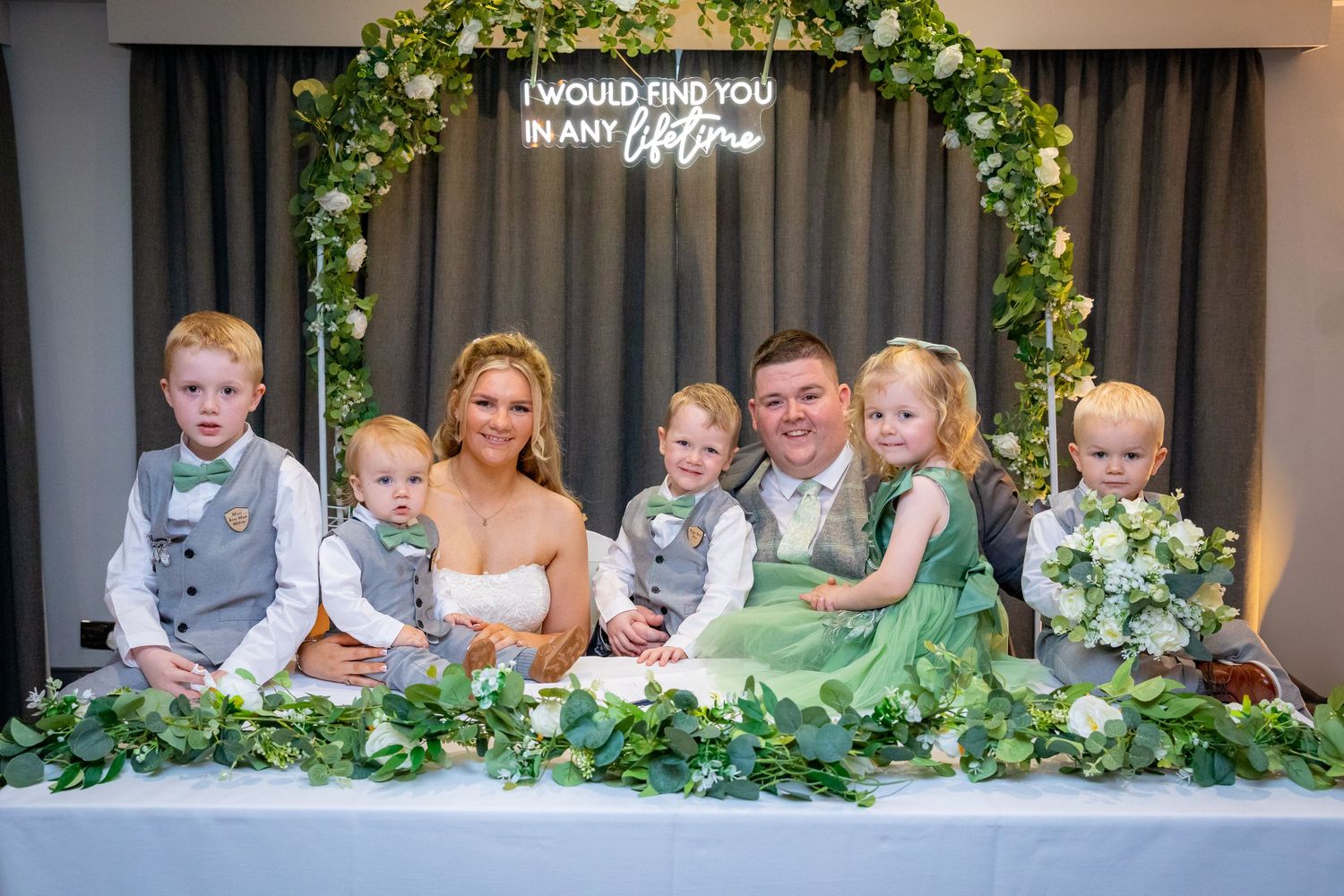Bride and groom pose with children at wedding reception table with floral arch and neon sign.