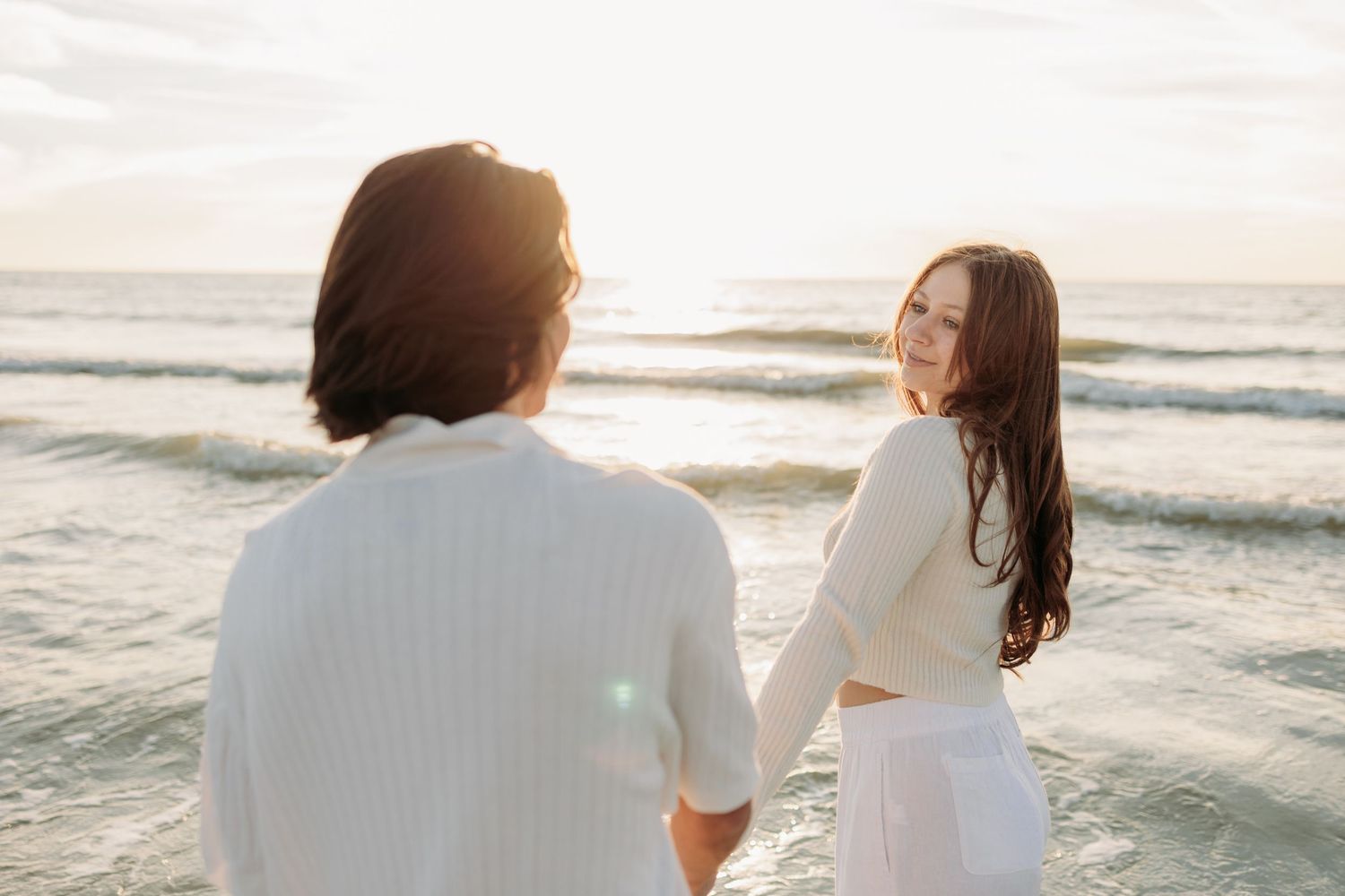 Couple holding hands and dancing on a sandy beach at sunset in white outfits with ocean waves behind them.