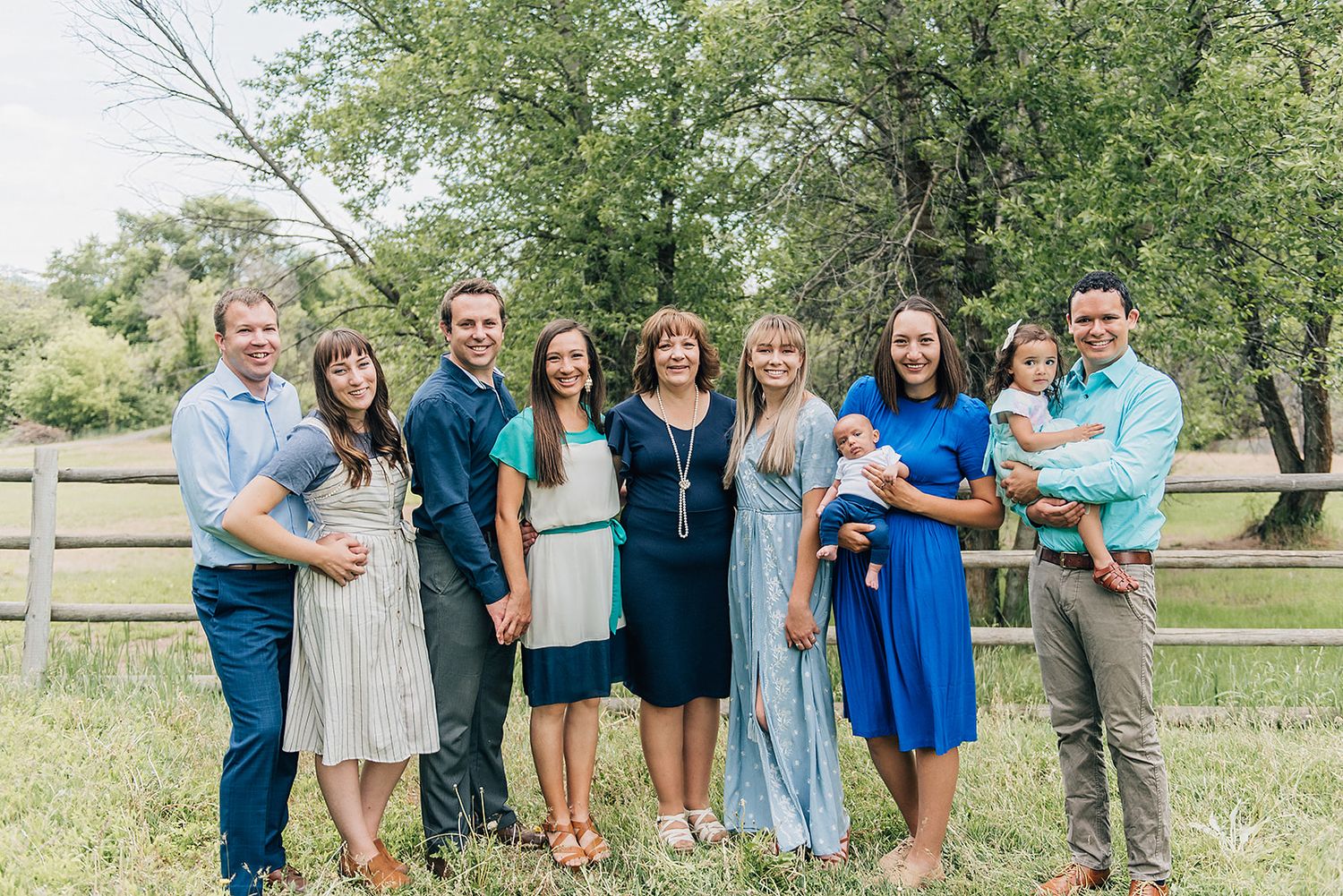 Group standing together outdoors in casual attire against a rustic fence and green foliage background.
