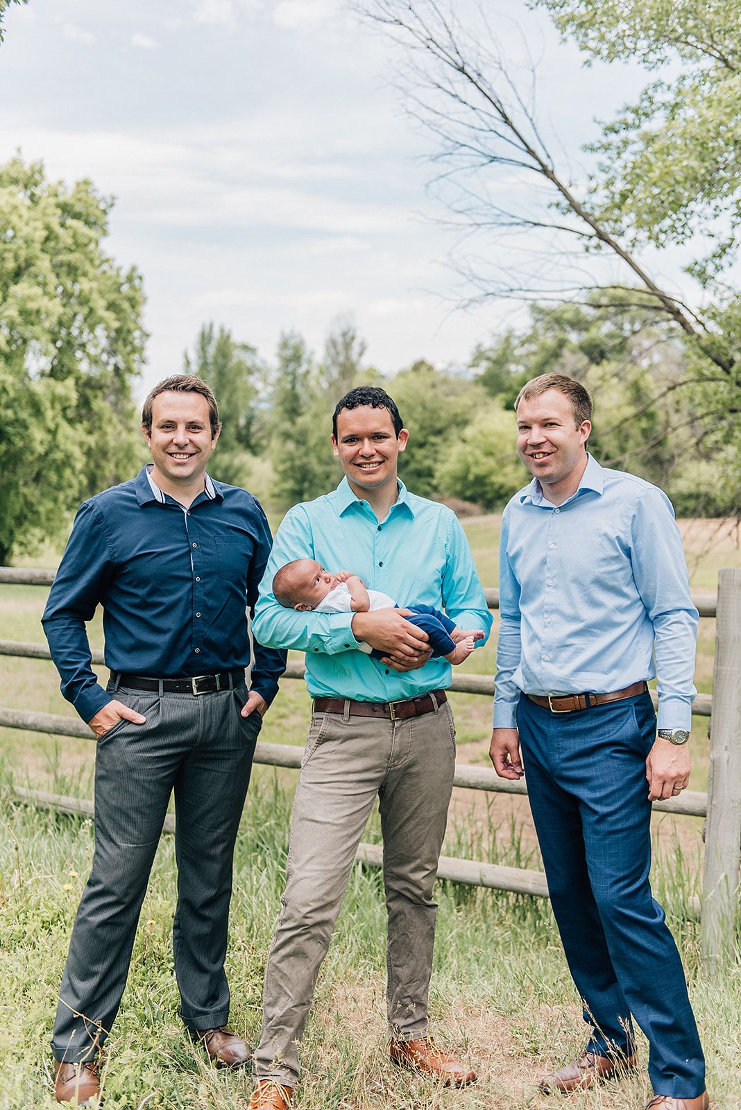 Three men in business casual attire pose together by a wooden fence in a pastoral setting.