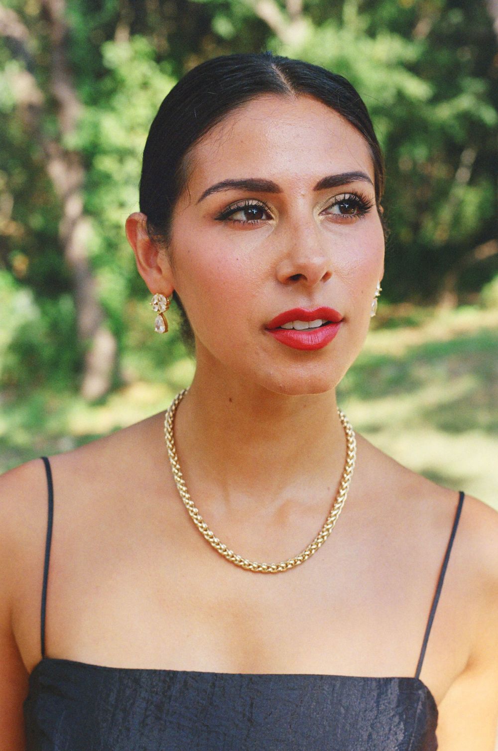 Woman wearing a gold chain necklace and black top poses against a blurred green nature background.