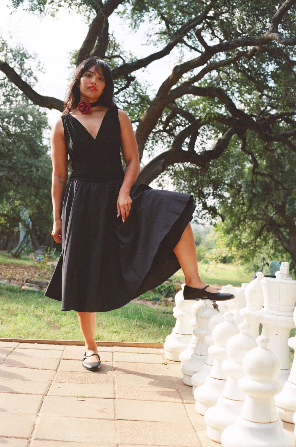 Woman in black dress poses beside white chess pieces on patio in outdoor garden setting.