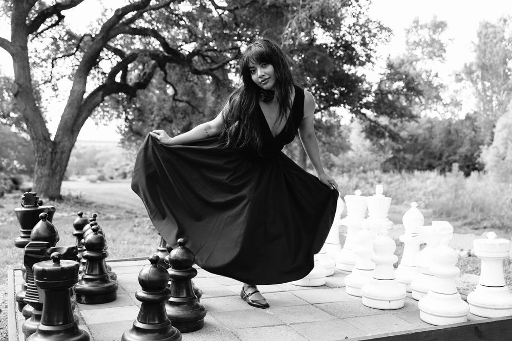 Black and white portrait with figure in elegant dress posed among large chess pieces outdoors.