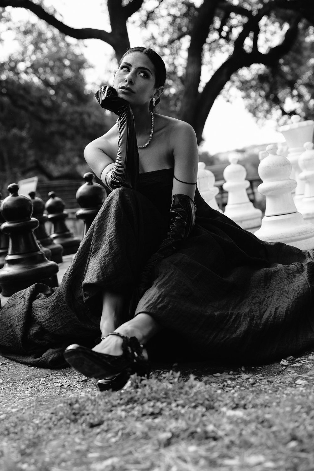 Dramatic black and white fashion photo with model posed elegantly among giant chess pieces.