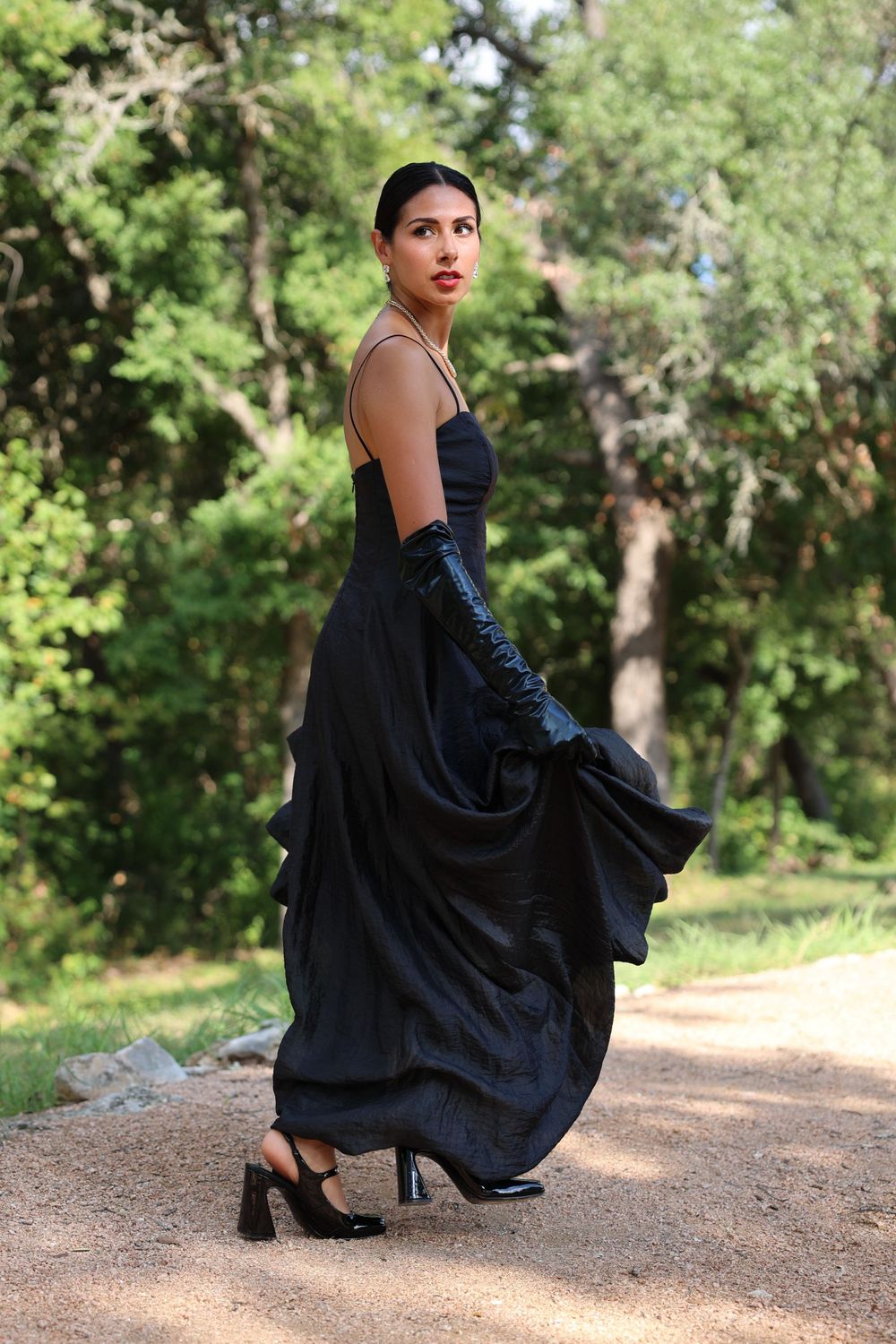 Elegant portrait of a figure in a flowing black dress and gloves in a garden setting.