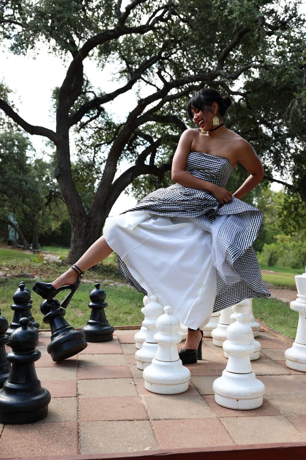 Person in checkered dress poses dramatically on giant chess board with large chess pieces.
