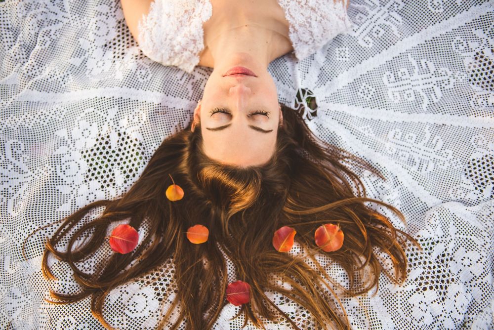 Woman with long brown hair lying on lace fabric with eyes closed, surrounded by colorful flower petals.