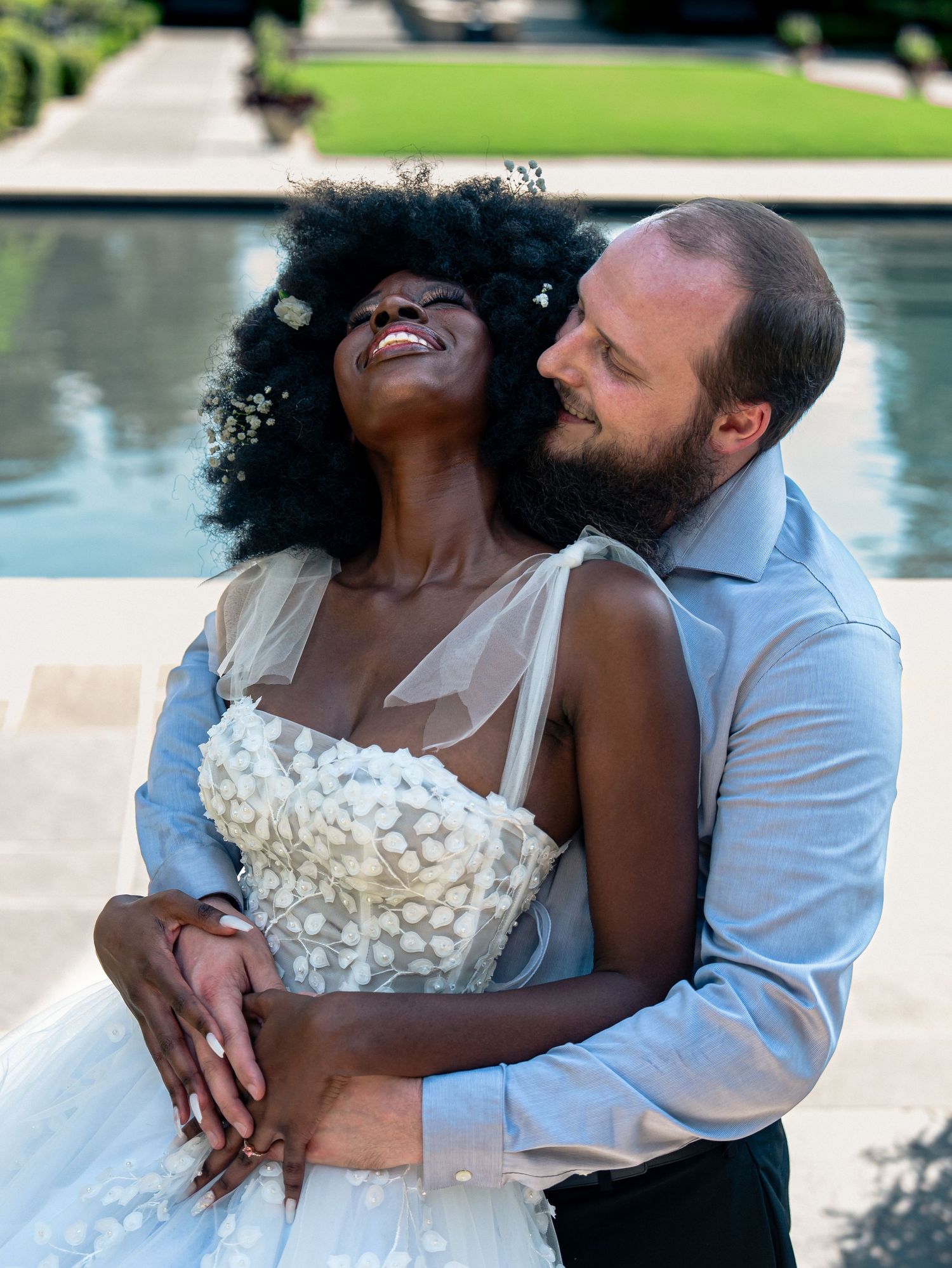 A romantic moment between a couple in wedding attire by a pool on a sunny day.