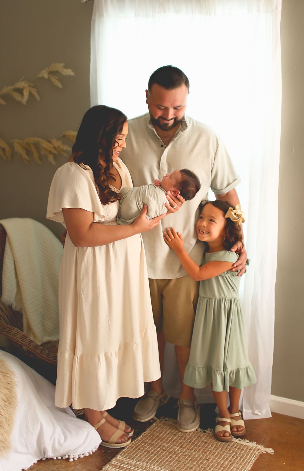 A family stands together near a window in a sunlit room wearing neutral tones and embracing a newborn baby.