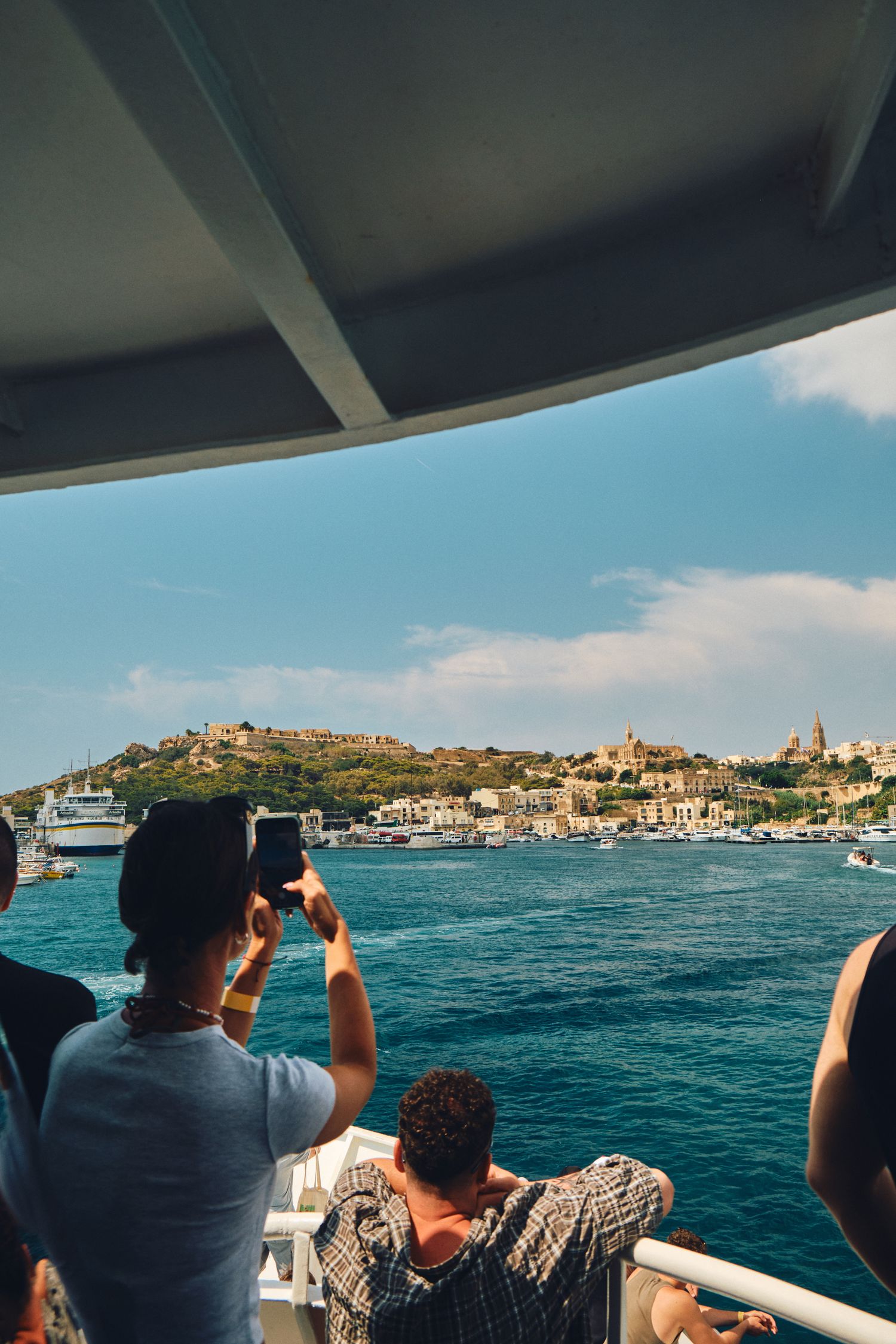 Tourists photographing Malta's coastal town and harbor from boat deck perspective.