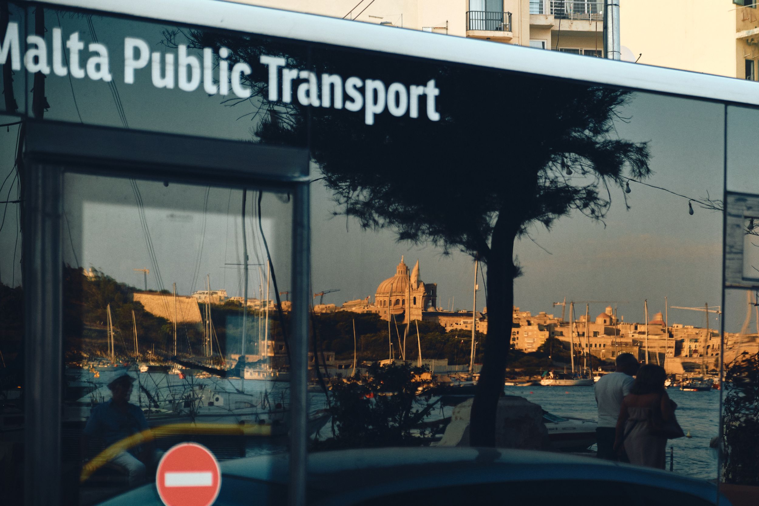 A Malta Public Transport bus window reflecting golden hour views of historic buildings and architecture.