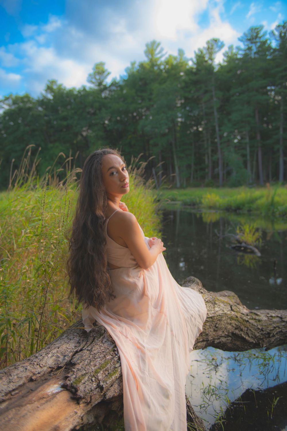 Someone in a flowing pink dress sits on a fallen log by a serene pond at sunset.