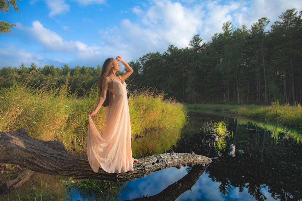 A flowing dress catches the breeze as someone poses on a log over a reflective pond.