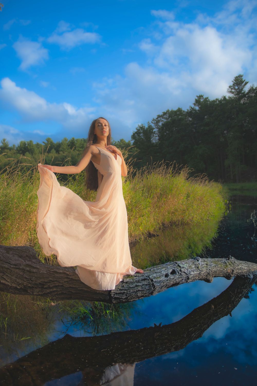 Someone in a flowing beige dress strikes a graceful pose on a fallen tree by a tranquil lake.