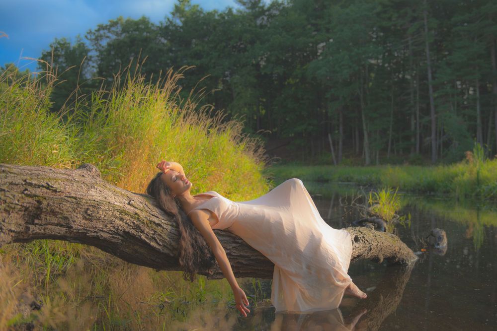 A dreamy summer scene shows someone in a light dress reclining on a log over water.